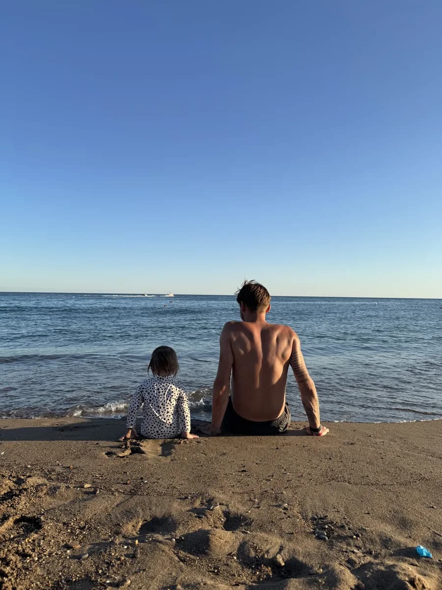 Vater und Kleinkind sitzen nebeneinander am Strand und blicken aufs Meer