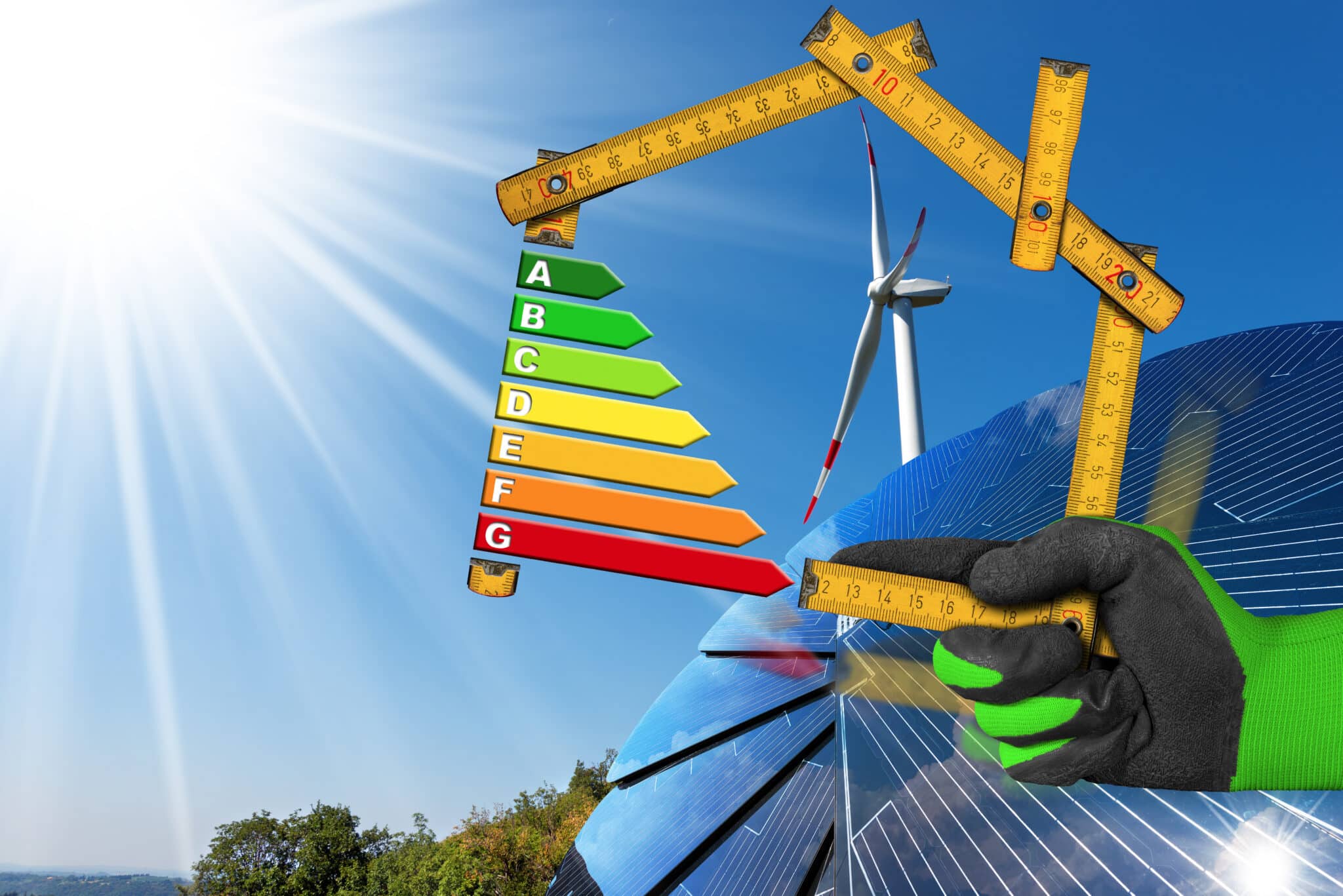 Hand with work glove holding a wooden folding ruler in the shape of house with energy efficiency rating on a blue sky with a solar panel and wind turbine